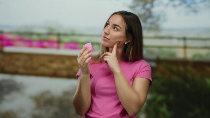Fototapeta premium Woman wearing pink shirt holds menstrual cup with pensive expression outdoors with blurred natural background suggesting feminine health awareness in casual setting.