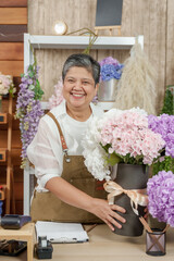 Asian senior female florist laughing while working at flower shop counter, arranging hydrangea...