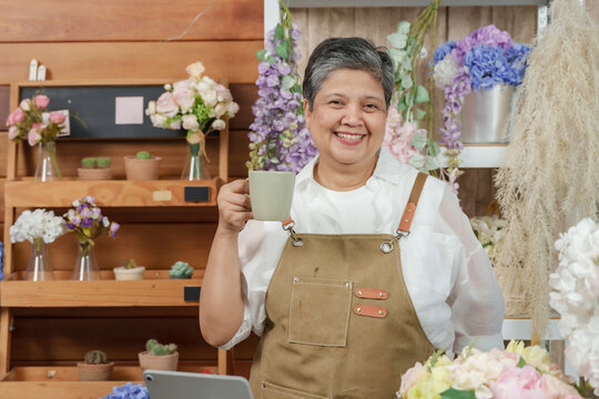 Asian senior female florist holding green ceramic mug while standing in flower shop full of bouquets and decorations, enjoying relaxing coffee break during online business working hours