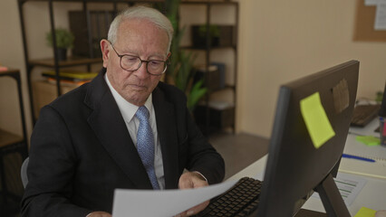 Senior man in a business suit working at a computer in a modern office interior, focused on reading documents and organizing tasks.