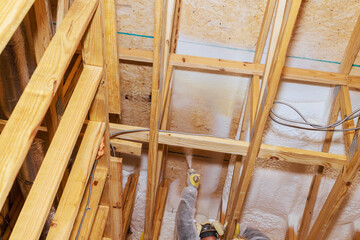 Skilled worker after install wooden beams, applies foam insulation material on ceiling framework in new house.