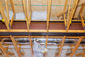 Worker in protective gear installs spray foam insulation with in exposed thermal barrier wooden...