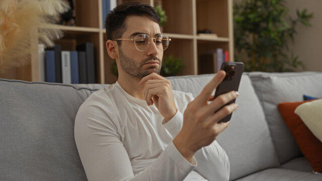 Young man in glasses sitting on couch in living room using smartphone with thoughtful expression surrounded by books and plants highlighting relaxed home environment