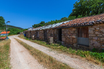 A rustic stone sheepfold with a red-tiled roof stands along a dirt road, surrounded by greenery and under a bright blue sky