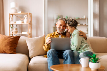Obraz premium Romantic happy senior spouses resting with laptop on couch in living room, embracing and using computer, browsing internet or video calling with family, free space