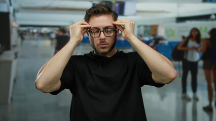 Young man with beard wearing glasses gesturing in an indoor airport terminal, surrounded by...