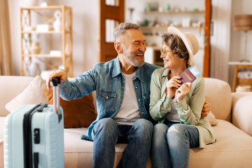 Joyful retired senior couple ready for trip, sitting on sofa at home, looking and smiling at each...