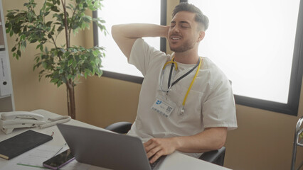Young male doctor with a beard in a hospital clinic wearing a stethoscope works on a laptop at his...