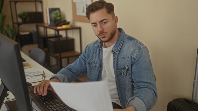 Young man with beard in denim jacket focused on work at office desk, reviewing documents by computer in modern workplace interior with shelves and plants.