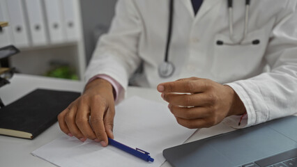 Young man in clinic wearing stethoscope writes notes at desk with hands visible, implying a medical...