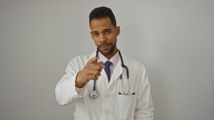 Confident young hispanic male doctor pointing at camera in white coat with stethoscope over...