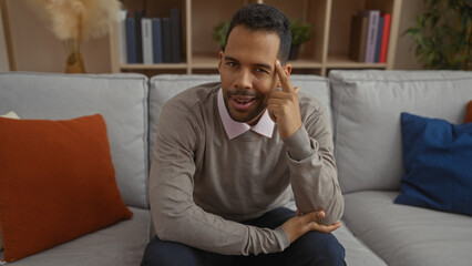 Young hispanic man sits confidently in a cozy apartment living room, exuding charm and...