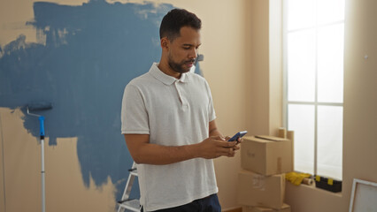 Young man in new home checking phone while renovating, featuring paint supplies in a sunlit living...