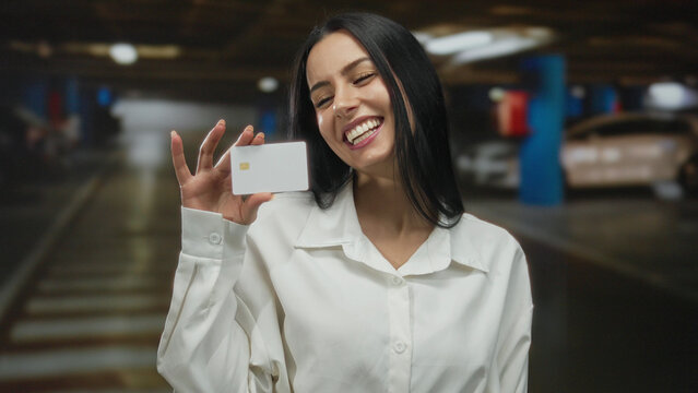 Young hispanic woman in parking garage smiling while holding credit card wearing white shirt indoors.