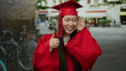 Woman in red graduation gown listening with hand to ear, standing on an urban street with bicycles...