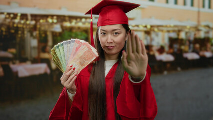 Woman wearing red graduation gown holding philippine peso banknotes outdoors in market area with a...