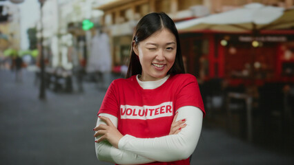 Woman smiling confidently on urban street wearing red volunteer shirt with blurred city background,...