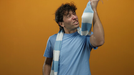Hispanic man cheering energetically with blue scarf against vibrant orange background showing...