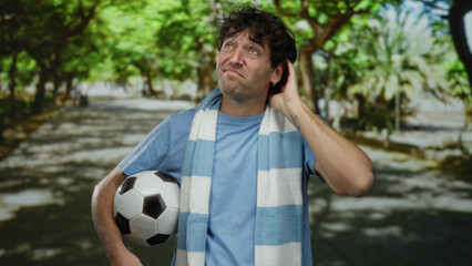 Hispanic man with a football at a park expresses uncertainty while standing on a sunny green...