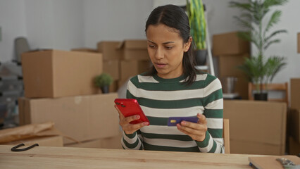 Woman using smartphone and credit card sitting indoors surrounded by moving boxes in her new...