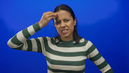 Young hispanic woman making a thoughtful expression against a blue background, wearing a striped...