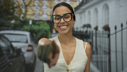 Young hispanic woman laughing on a city street outdoors, smiling brightly with glasses and wearing a casual top while pointing playfully at the camera.