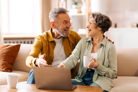 Tech-savvy seniors. Happy elderly spouses using laptop and chatting, watching funny content, relaxing on sofa in living room