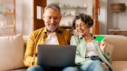 Happy modern senior spouses shopping online via laptop with credit card, man and woman making...