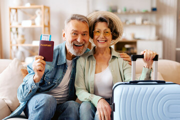 Excited senior couple ready for new trip, holding travel tickets and passports, sitting with packed...