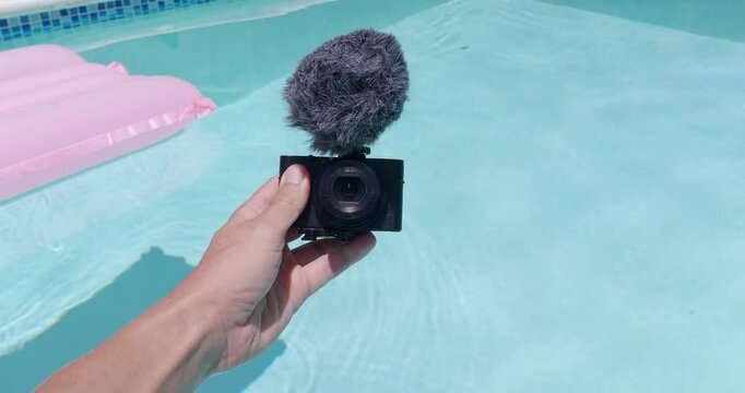 POV. Hand holding a compact camera with microphone above a swimming pool during a hot sunny day. Vlog concept. 