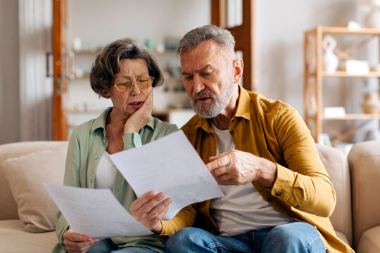 Elderly spouses checking financial documents, calculating family budget, sitting on couch in living room at home