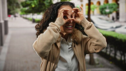 Woman with curly hair playfully mimics binoculars on a city street, exuding happiness and curiosity...