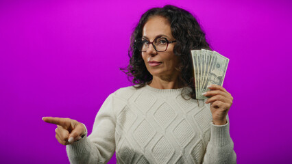 Middle-aged woman holding american dollars against an isolated purple background, wearing glasses...