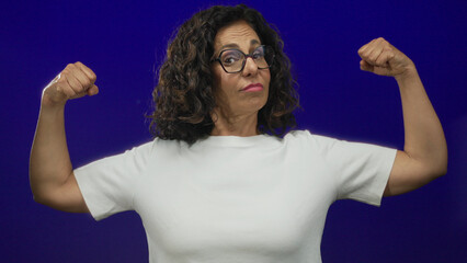 Hispanic middle-aged woman confidently flexing her muscles against an isolated blue background...