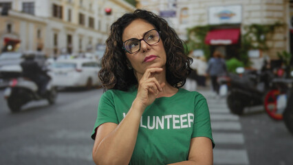 Woman in green volunteer shirt standing thoughtfully on a busy street with cars and buildings in...