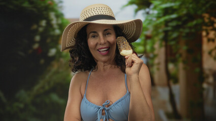 Hispanic woman in a bikini enjoys an ice cream outdoors in a green park, wearing a sun hat and...