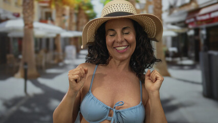 Middle-aged hispanic woman in bikini celebrates joyfully on a lively street, wearing a sun hat,...