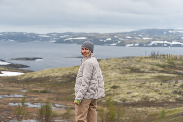 Fototapeta premium Smiling woman in beige jacket and gray beanie standing on Arctic tundra with Barents Sea and snow-covered hills in background