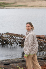 Woman in a beige puffer jacket and sunglasses standing near rusted shipwrecks by the shore, looking...