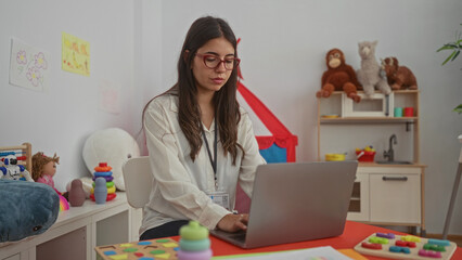 Young hispanic woman teacher working indoors on a laptop in a kindergarten surrounded by...
