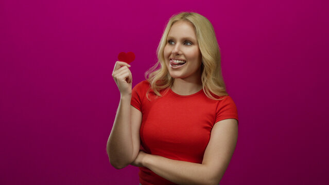 Young blonde woman holding heart with playful expression against pink background wearing red shirt