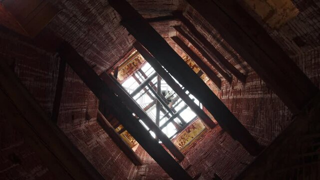 Large wooden beams barely support walls of tall bell tower of brick temple, which has collapsed and leaky roof, through which sky is visible. rotating camera. Bottom view