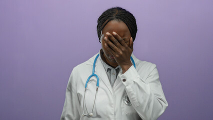 Female doctor with braided hair stands in front of a purple wall covering her face with her hand,...