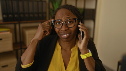 Woman talking on phone in office wearing glasses and yellow shirt displaying thoughtful expression...