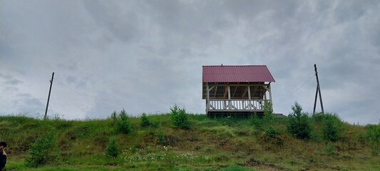 A small house situated on a hill surrounded by lush green grass and plants, under a clear blue sky adorned with white clouds. The landscape captures the essence of a peaceful rural area.