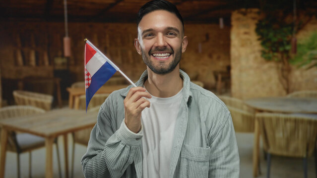 Young man holding croatian flag smiling on restaurant terrace with tables in the background outdoors.