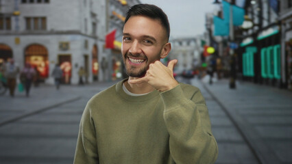 Fototapeta premium Young man on busy city street gestures a phone call with a playful smile, showcasing urban lifestyle and communication in an outdoor urban environment.