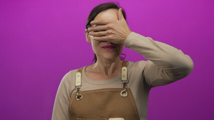 Woman in aprons smiling playfully covering eyes against vibrant pink background showcasing colorful isolated scene with cheerful expression.