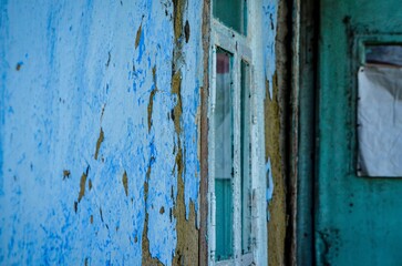 A blue wall with peeling paint and a window with a white frame. The window is dirty and the frame is chipped
