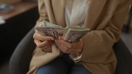 Woman counting stack of us dollar bills in modern office setting, hands focused, wearing beige blazer, highlighting financial themes in a professional indoor environment.